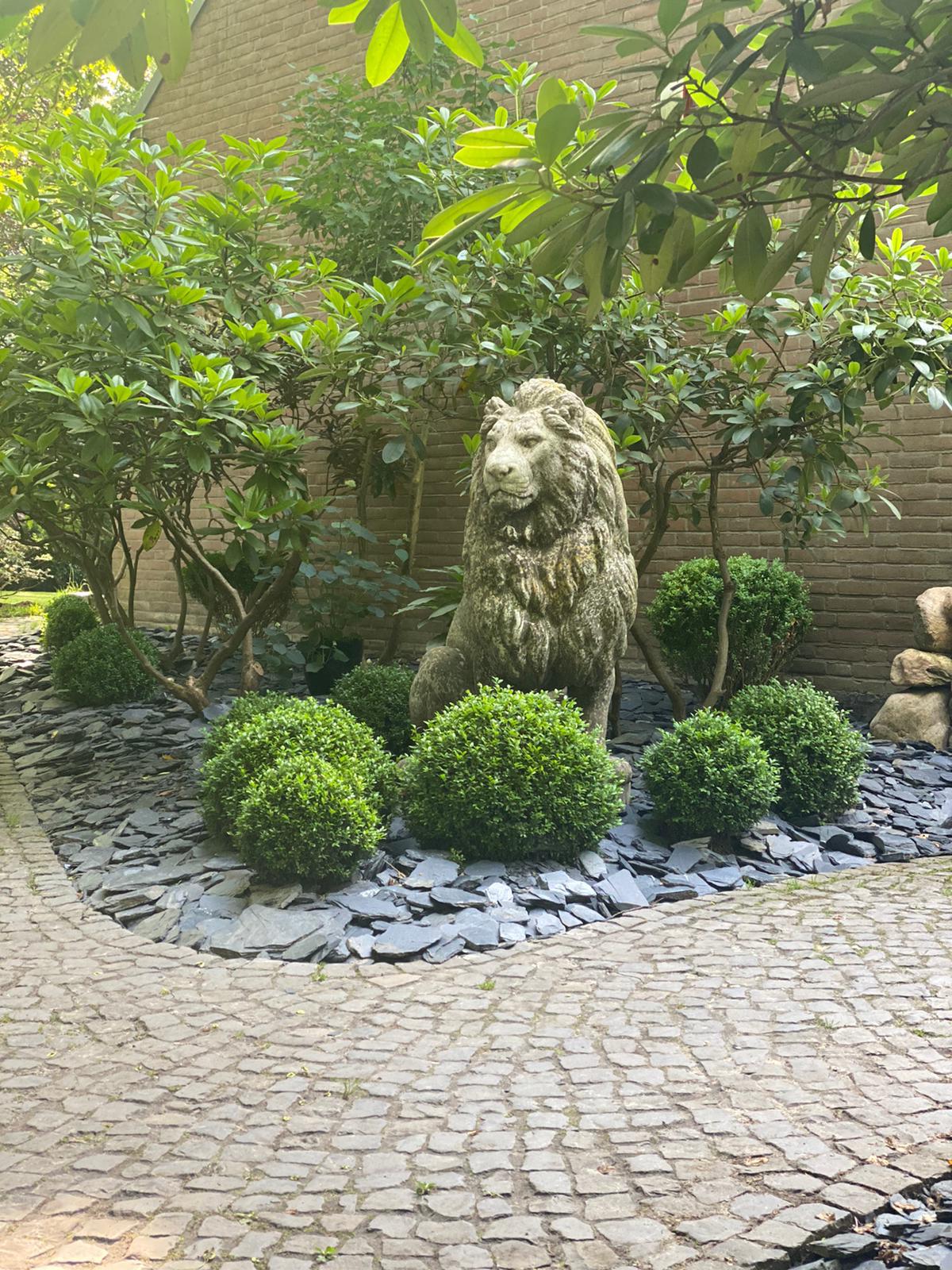 Ein gepflasterter Weg, der durch einen Garten führt, mit einer majestätischen Löwenstatue in der Mitte.